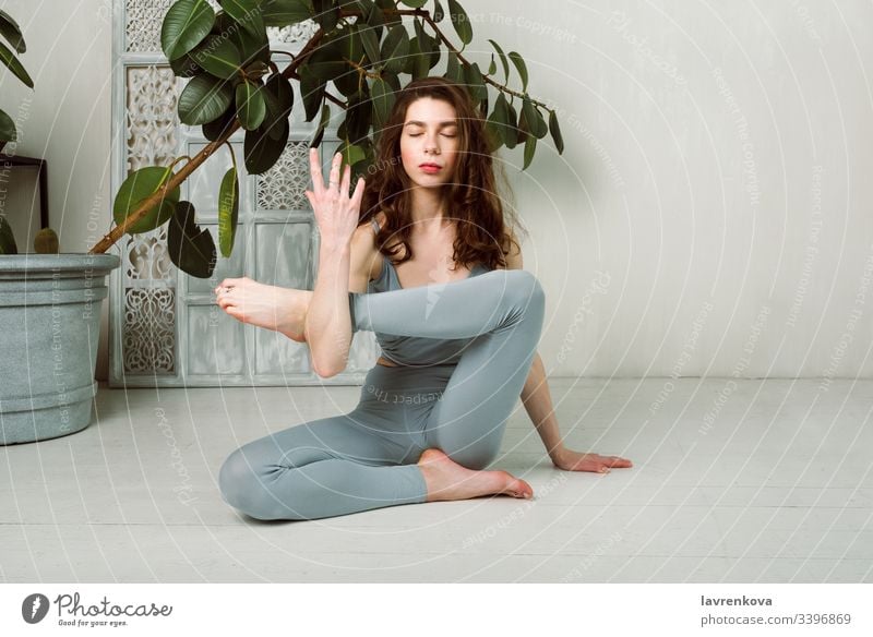 Young yogini practicing on the floor in a room with white walls and plant workout active sport exercise skinny slim leggings brunette calm flexible meditation
