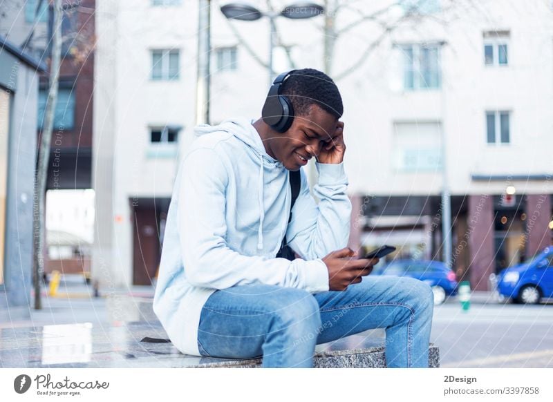 Young Black man sitting on bench while listening music by headphones technology lifestyle male black casual attire trendy happy african earphones guy people