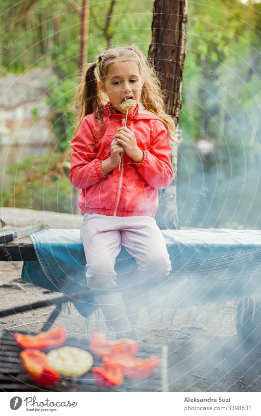 Man and his little daughter having barbecue in forest on rocky shore of lake, making a fire, grilling bread, vegetables and marshmallow. Family exploring Finland. Scandinavian summer landscape.