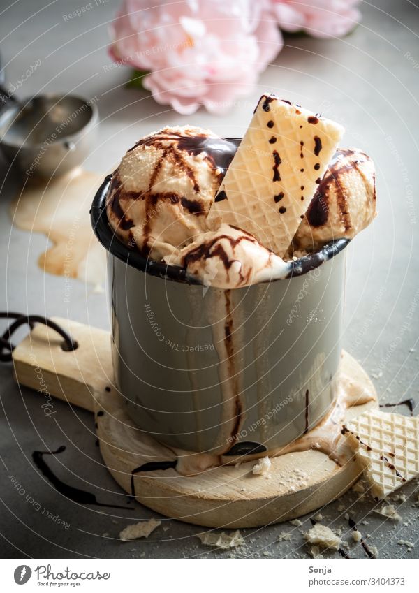 Geschmolzenes Vanilleeis mit einer Eiswaffel in einem rustikalen Becher und rosa Blüten auf einem grauen Hintergrund vanilleeis eiswaffel schokoladesauce