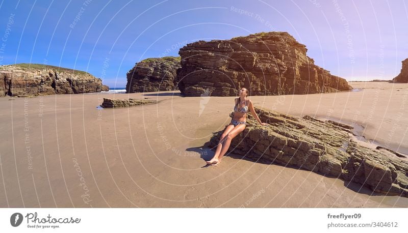 Young woman in bikini sitting on a rock on a beach full of cliffs tourism hiking galicia spain ribadeo castros illas atlantic bay touristic cathedrals ocean