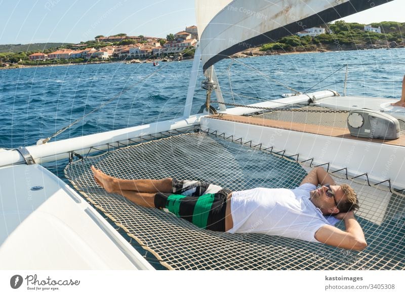 Sporty man relaxing, lying in hammock of a catamaran sailing boat on luxury nautic vacations near picture perfect Palau town, Sardinia, Italy. sea sailingboat