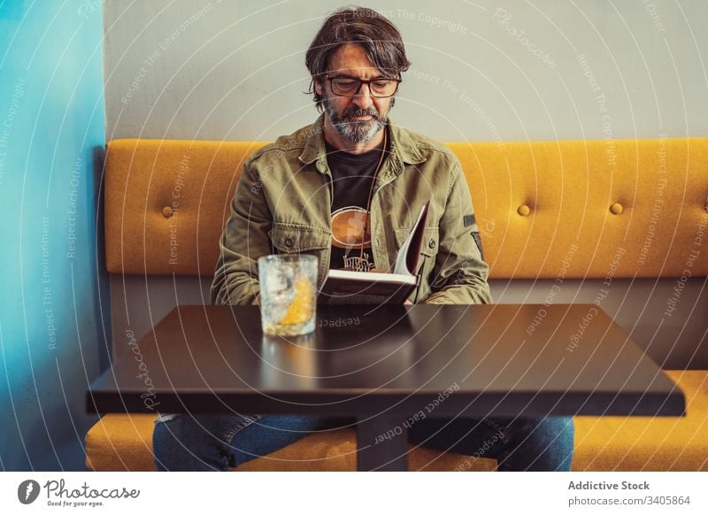 Bearded man reading book in cafe sofa table cocktail serious sit male casual modern lifestyle mature adult relax rest bar drink beverage smart glasses guy story