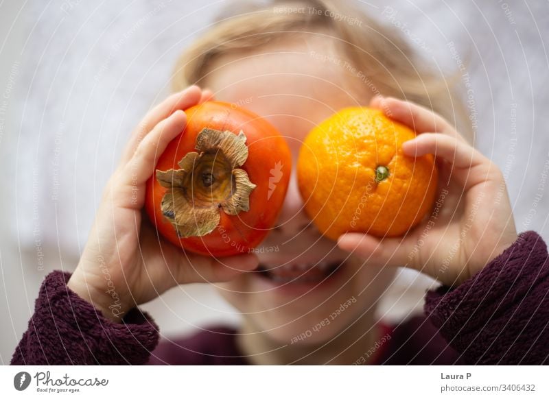 Little girl holding a kiwi and an orange in front of her eyes, laughing cute little child kid playing fun smiling fruits hands hiding hiding behind