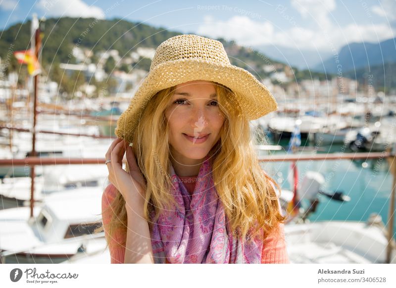 Portrait of young woman with light curly hair in straw hat enjoying sun and breeze, smiling. Sunny harbor with boats and yachts, green mountains on background. Enjoying life, happy person traveling,