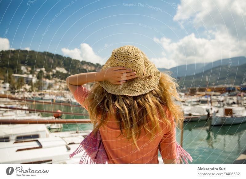 Portrait of woman with light curly hair, rear view,  holding straw hat with hand. Sunny harbor, boats and yachts, green mountains on background. Enjoying life, happy traveling,
