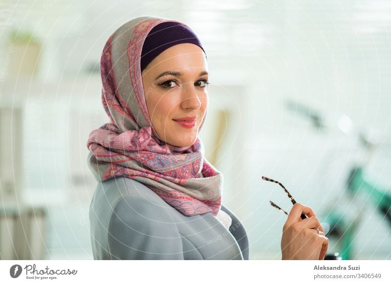 Beautiful stylish woman in hijab and eyeglasses, sitting at desk with laptop in office. Portrait of confident muslim businesswoman. Modern office with big window, bicycle on background.