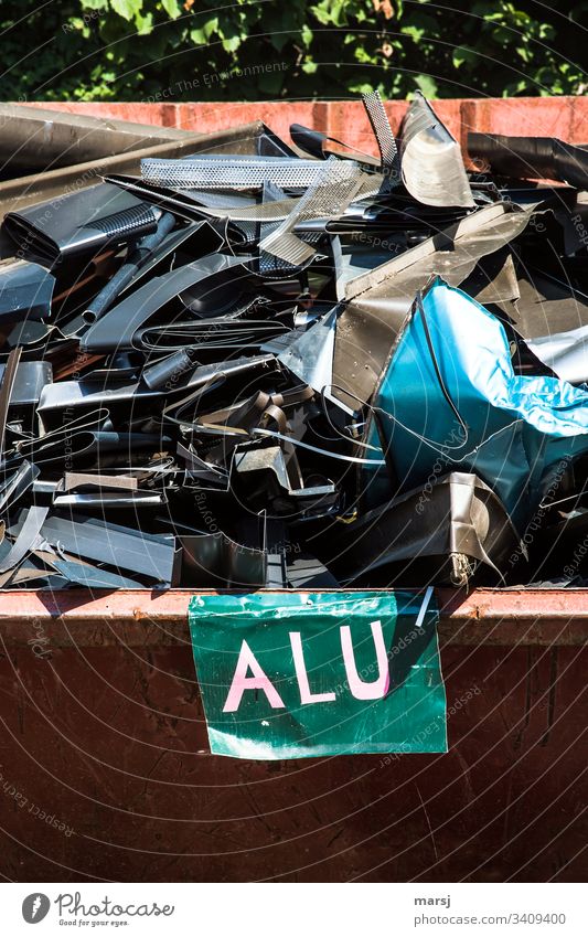 A scrap container full of, partly coloured, aluminium scrap from a roofer. In the front a board with the inscription Alu Scrap metal Aluminium variegated