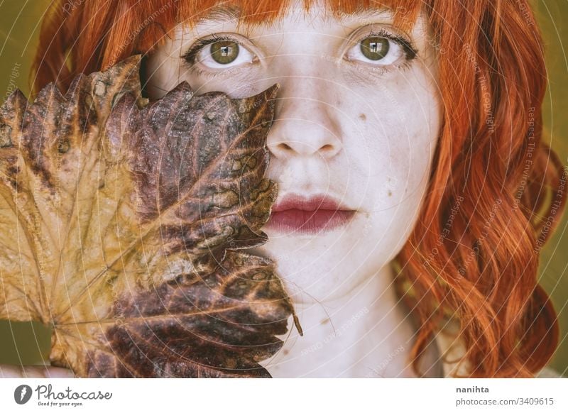 Redhead young woman covering by a dry autumn leaf fall leave portrait art artistic redhead red hair delicate face ginger concept conceptual youth orange yellow