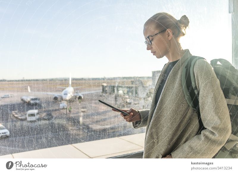 Casually dressed female traveler at airport looking at smart phone device in front of airport gate windows overlooking planes on airport runway woman girl
