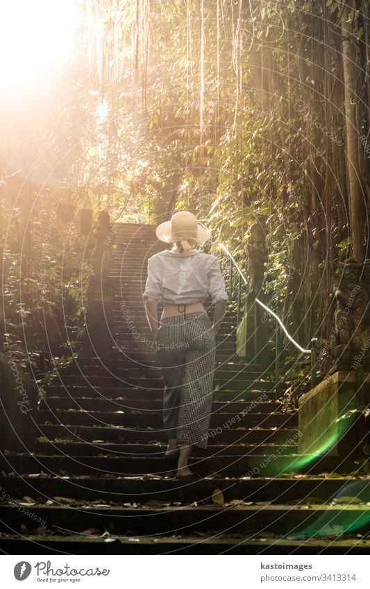 Rear view of beautiful fashionable stylish female traveler wearing a hat, walking the staircase of traditional hidu tample around Ubud on Bali. Concept of luxury vacation, exploring and discovering the world.