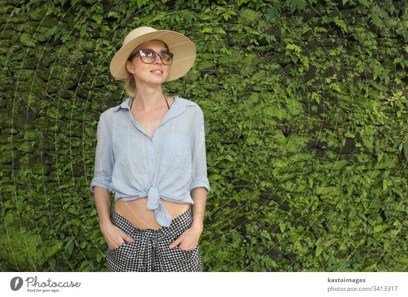Portrait of a beautiful female traveler. Smiling young woman in summer hat wearing sunglasses, standing in front of lush tropical plant greenery wall background.