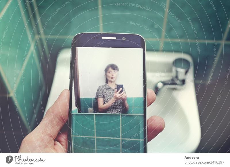 In front of a sink in a turquoise tiled bathroom, a left hand holds a mobile phone with a selfie of a woman smartphone Selfie photo Woman tiles Sink by hand