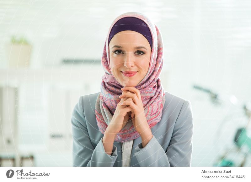 Beautiful stylish woman in hijab and eyeglasses, sitting at desk with laptop in office. Portrait of confident muslim businesswoman. Modern office with big window, bicycle on background.