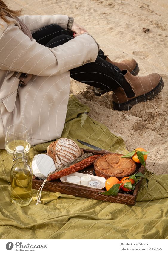 Crop woman sitting near wine and tray with pastry and tangerines with sausages and cheese picnic beach food blanket rest sand female bread pie drink beverage