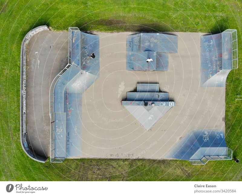 Aerial view  of a skatepark with 2 unidentifiable teens. Grey-blue ramps against a grass background.  created by dji camera Skatepark skateboarding scooters