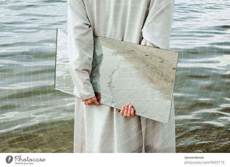 Closeup of woman standind on a shore in blue dress holding a mirror with beach reflecting in it, selective adult caucasian clean coast concept environment