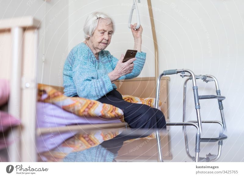 Elderly 96 years old woman reading phone message while sitting on medical bed supporting her by holder. senior home smart technology smartphone mobile
