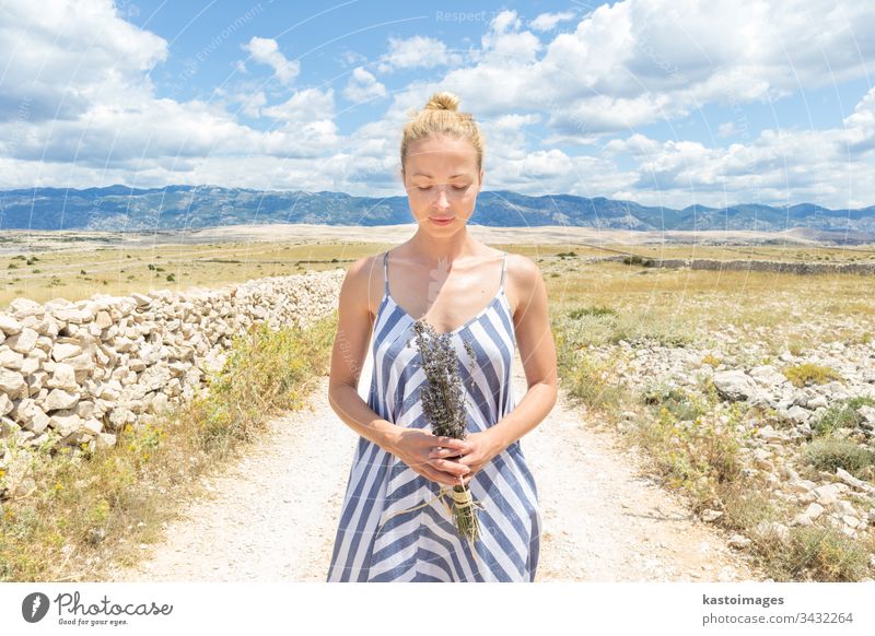 Caucasian young woman in summer dress holding bouquet of lavender flowers enjoying pure Mediterranean nature at rocky Croatian coast lanscape on Pag island in summertime