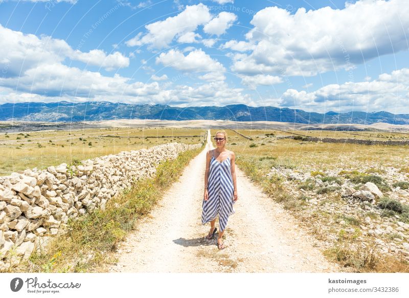 Caucasian young woman in summer dress holding bouquet of lavender flowers while walking outdoor through dry rocky Mediterranean Croatian coast lanscape on Pag island in summertime