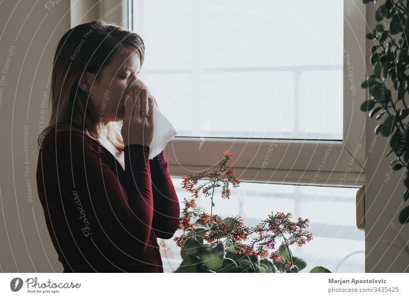 Young sick woman blowing her nose into a handkerchief by the window 30s adult allergy alone apartment care casual caucasian cold contagion dark disease epidemic