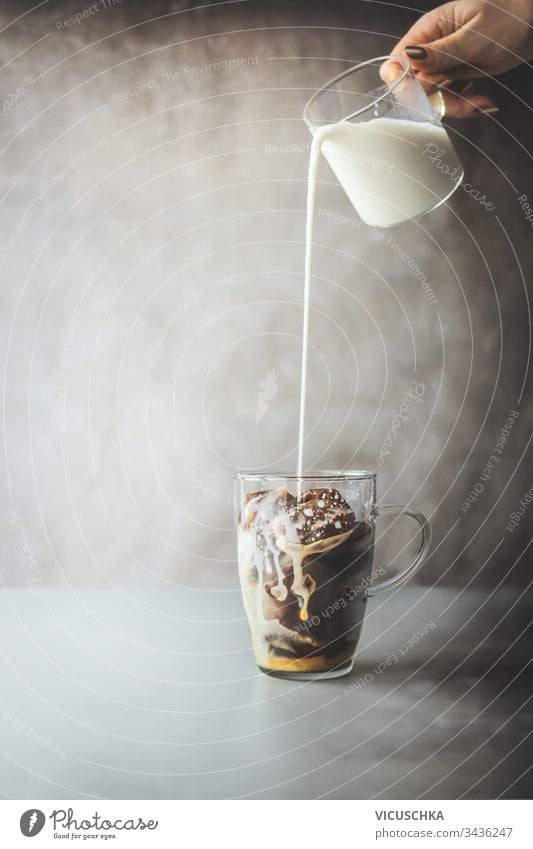Ice coffee preparation. Female hand pouring cream milk in glass with coffee ice cubes on rustic table at concrete wall background. Iced coffee making. Summer refreshing beverage. Cold drink.