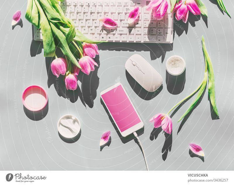 Female desktop with light pink tulips flowers bunch, cosmetics,  PC keyboard , smartphone with blank screen and wire . Top view. Mothers day . Springtime. Greeting