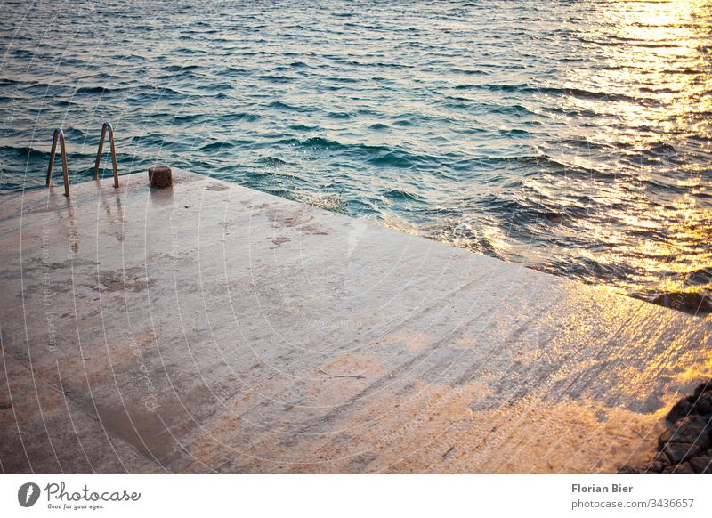Wet concrete sunbathing area with swimming ladder into the sea at sunset Concrete Concrete floor Ladder lying surface Ocean Waves Sunset reflection Water