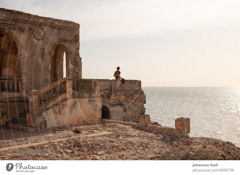 Young woman sitting on the parapet of a ruin and looking at the sea Ocean ocean Water Beach Coast Ruin Debris stones rock masonry Architecture Temple castle