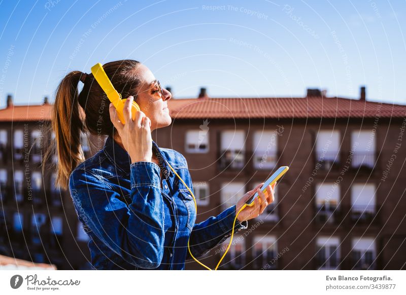 young woman listening to music on mobile phone and yellow headset. Fun and lifestyle technology fun terrace outdoors sunny sunglasses happy caucasian active