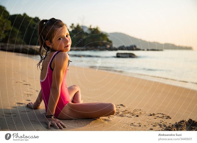 Cute happy little girl playing with sand on the beach in swimming suit, drawing a heart and writing. Beautiful summer sunset, sea, coconut palms, picturesque exotic landscape. Phuket, Thailand