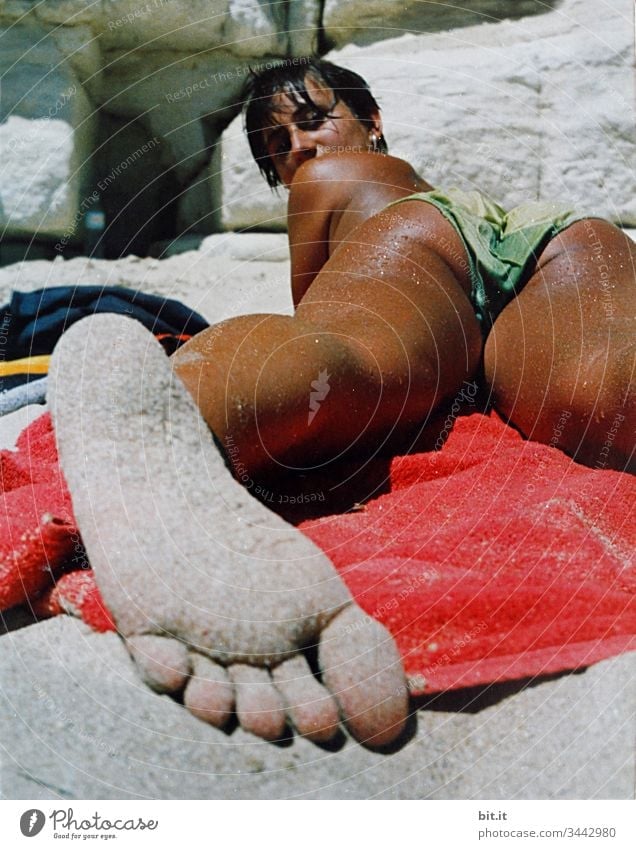 Young woman lies tanned on the beach on a towel, barefoot with sandy feet in the foreground and looks into the camera. Beach Woman Brown Sunbathing Summer
