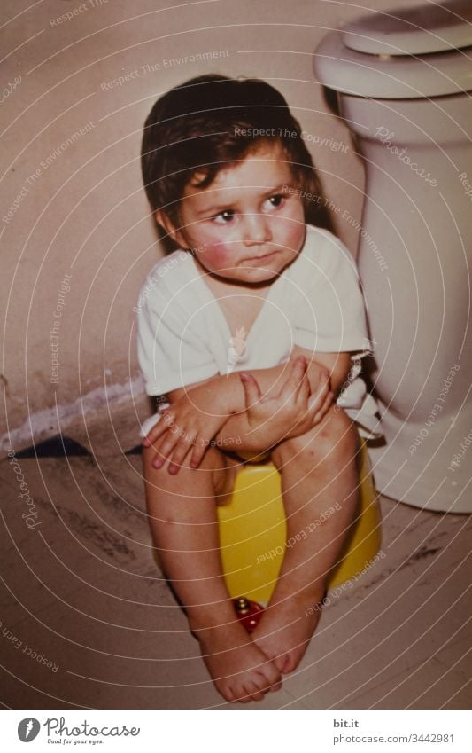 Pondering the toilet paper situation, the little girl sits on the potty in the bathroom in front of the toilet, with a bed bottle on her bare feet and arms crossed.