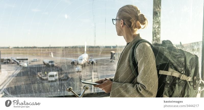 Young woman standing near airport gates window holding cellphone in her hands, wearing travel backpack and walking to lounge area. Female traveler search online map at the airport