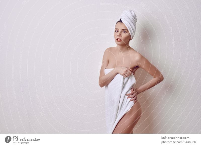 Alluring young naked woman covering her breasts and front of her torso with a fresh clean white a towel as she looks at camera with a sultry expression and parted lips over a white studio background