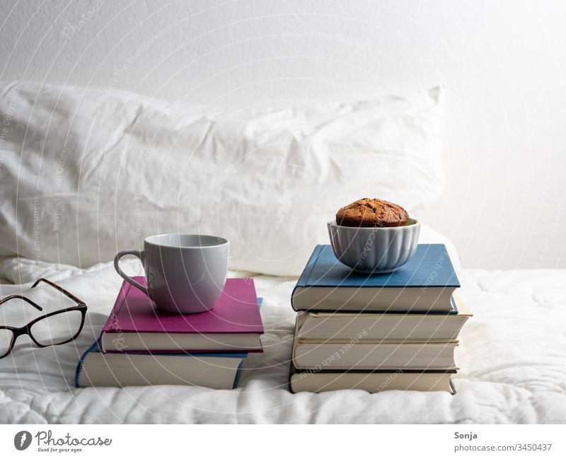 Bücherstapel mit einer Tasse Kaffee und einem Muffin auf einer weißen Bettdecke im Schlafzimmer bücherstapel muffin hygge bettwäsche schlafzimmer Isolated