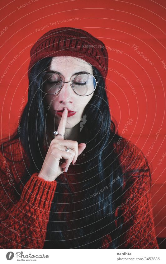 Young woman asking for silence against a red background calm gesture quiet tranquility young nerd nerdy glasses portrait portraiture studio brunette hand cover