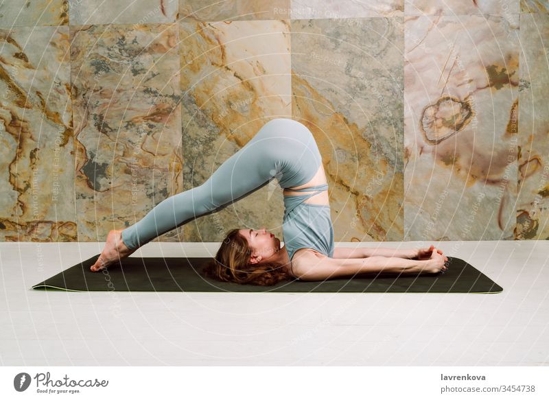 Young yogini practicing Halasana (plow pose) in pastel blue leggins and top, selective focus active athlete athletic attractive body brunette calm caucasian