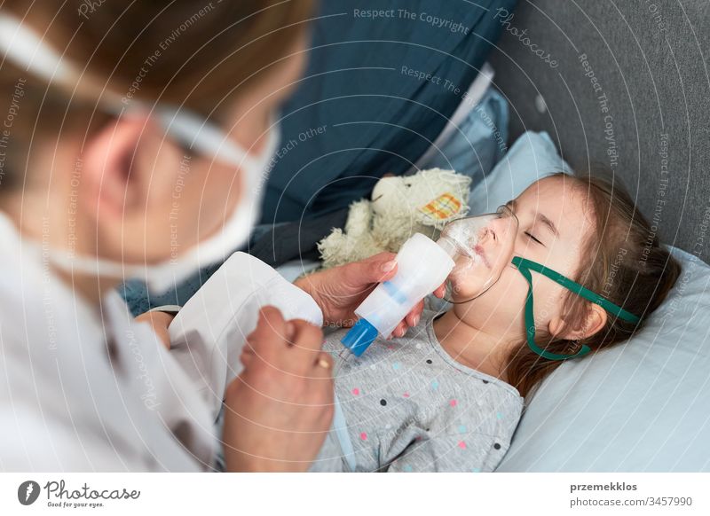 Doctor visiting little patient at home. Child having medical inhalation treatment with nebuliser. Girl with breathing mask on her face. Woman wearing uniform and face mask