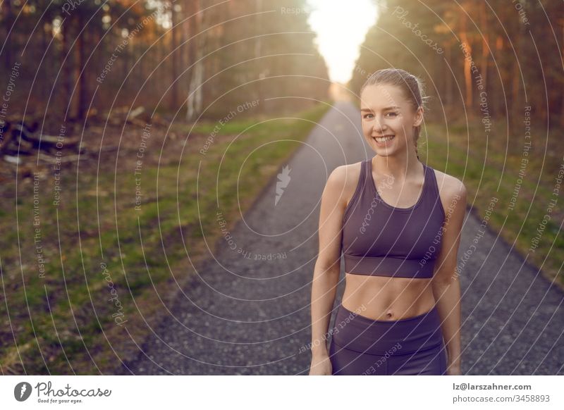 Smiling athletic fit young woman working out on a tarred lane through forests backlit by the warm glow of the sun in a healthy active lifestyle concept face