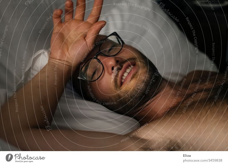 A man with a beard and glasses, who lies comfortably and sleepily in bed and touches his forehead with his hand Man Bed Eyeglasses Facial hair Forehead Hand
