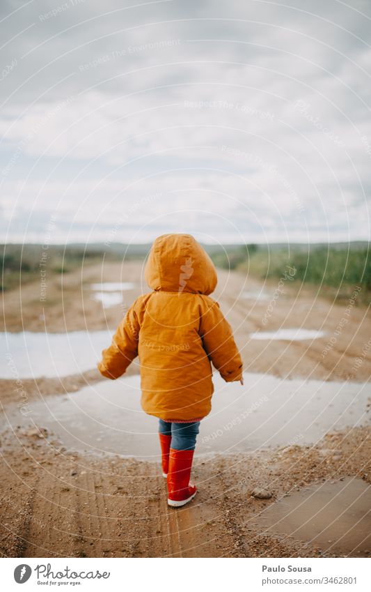 Rearview child walking in a puddle with orange jacket Orange Rubber boots Red Puddle Wet Exterior shot Colour photo Rain Human being Joy Child Water Playing