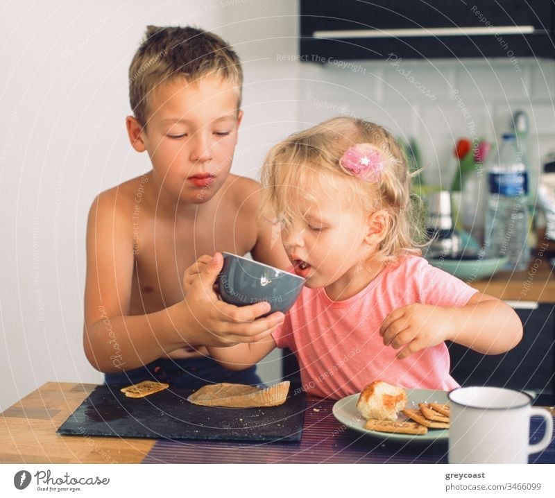 Elder brother taking care of junior sister and helping her drink from the cup during breakfast. Everyday morning moments children tea meal take care siblings