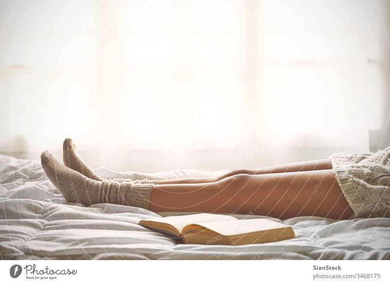 Soft photo of woman legs on the bed with old book next her read reading window view lazy sunday winter coffee cup tea morning girl home relax socks beautiful