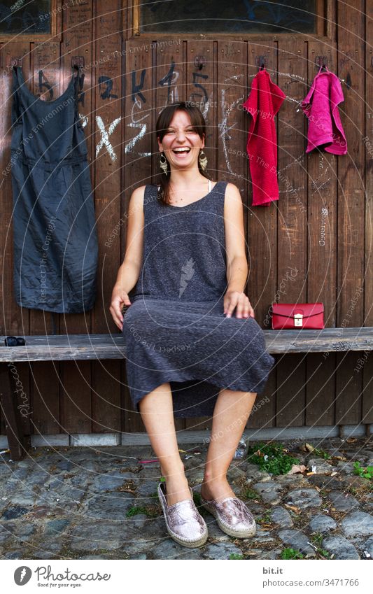 Young woman sits laughing on a bench, in front of a wooden wardrobe with a dress and towels hanging on it. Woman Youth (Young adults) girl Human being teenager
