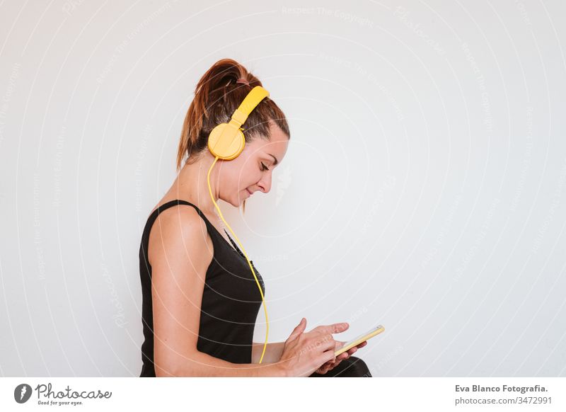 Young woman listening to music on yellow mobile phone and headset. Sitting on yoga mat, relaxing after exercise. Healthy lifestyle indoors technology internet