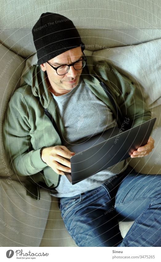 HomeOffice: relaxed man lying on the sofa with his notebook home office Workplace Notebook Sofa cap Hipster Jeans Eyeglasses Easygoing casually Man independent