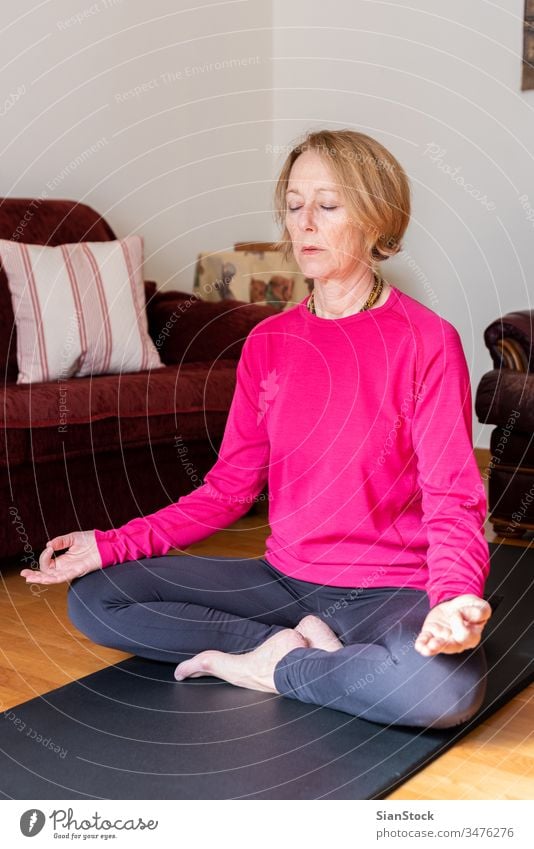 Middle aged woman sitting in lotus position on yoga mat in her living room. meditation lotus pose beauty workout european exercising flexibility wellness single