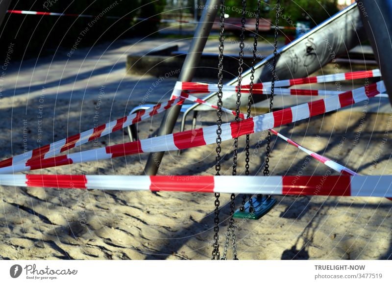 Corona Thoughts | No Playing! Red and white barrier tape criss-crossing a children's swing in a playground with sunlight and shade covid-19 Alarm