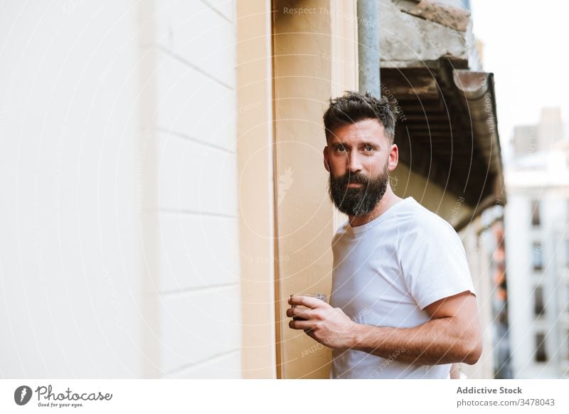 Bearded guy with hot drink resting on balcony man morning casual stand modern home apartment male beard adult handsome lifestyle relax early fresh beverage